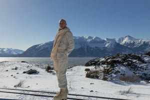 Musician Quinn Christopherson standing against the backdrop of snowy Alaskan mountains in an ALL WE REMEMBER undyed organic cotton scarf, flannel shirt, and twill pants.